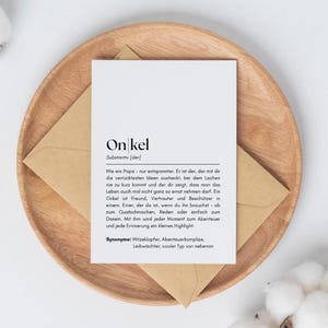 Könnte beinhalten: Eine weiße Karte mit dem deutschen Wort "Onkel" aufgedruckt, was "Onkel" bedeutet, mit einer Definition darunter. Die Karte liegt auf einem Holztablett mit einem braunen Umschlag. Die Karte ist von einem Wattebausch umgeben.