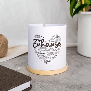 Pode incluir: Um castiçal cilíndrico branco com a palavra "Zuhause" em forma de coração, rodeado por palavras como "Leben" e "Glück". O suporte assenta numa base castanha clara. Uma bola de barbante e um caderno estão no fundo.