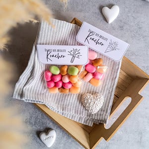 Op de afbeelding: Een kleine doorzichtige zak gevuld met kleurrijke snoepjes met een wit label waarop "Du bist der absolute Kracher" staat met een vuurwerk afbeelding. De zak ligt op een gestreepte grijs-witte stof met een houten dienblad eronder.