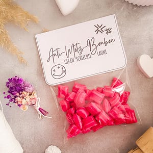 Könnte beinhalten: Ein klarer Beutel mit leuchtend rosa Bonbons, neben einer weißen Karte mit dem Text "Anti-Motz-Bonbon" und einem Smiley. Ein kleiner Strauß Trockenblumen und ein weißes Herz sind ebenfalls zu sehen.