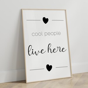 May include: Black and white print with the text "cool people live here" and two black hearts. The print is framed in a light wood frame.