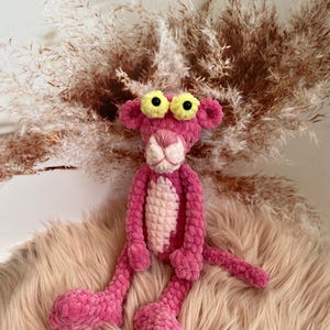 May include: Pink crocheted plush toy shaped like a panther with yellow eyes. The toy is sitting on a light brown fuzzy surface.