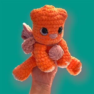 Puede incluir: Un juguete de peluche naranja y rosa tejido a crochet con ojos grandes y una concha en su mano. El juguete está sentado en una mano.