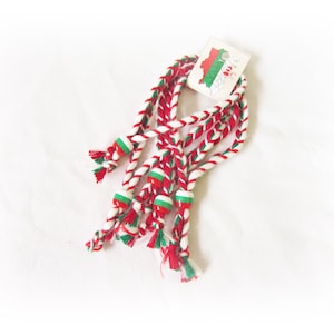 May include: Braided red, white, and green decorative cords with small red and green beads. A small tag with a map and flag design is attached to the cords. The cords are arranged on a white background.