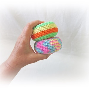 May include: Two colorful, handmade crochet juggling balls. The top ball features green, orange, and yellow stripes, while the bottom ball displays a mix of pink, blue, and yellow. The balls are held in a hand against a white background.