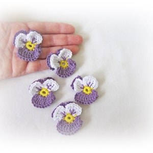 May include: Five crocheted purple and white pansies with yellow centres. The pansies are arranged on a white surface.