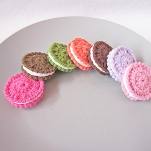 May include: A collection of colorful, crocheted macaron-shaped items arranged on a gray plate. The macarons are in various colors, including pink, green, orange, brown, and purple, with white filling. These handmade items are likely decorative or for play.