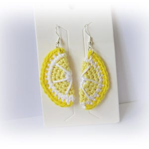May include: A pair of handmade crochet earrings in the shape of lemon slices. The earrings are made with bright yellow and white yarn, with silver-coloured ear wires. The earrings are displayed on a white card, a fun summer accessory.