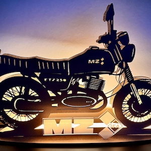 Könnte beinhalten: Eine Holz-Motorrad-Silhouette-Lampe mit dem Text "MZ ETZ 250" und "IFA" auf der Basis. Die Lampe hat einen warmen Schein, der die komplizierten Ausschnittdetails des Motorraddesigns hervorhebt. Der Hintergrund ist ein Farbverlauf aus Blau und Gelb.