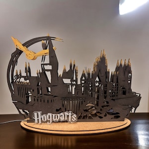 Könnte beinhalten: Eine schwarze und goldene Holzsilhouette von Hogwarts mit einem goldenen Schnatz, der darüber fliegt. Die Basis der Silhouette ist mit dem Wort "Hogwarts" beschriftet.