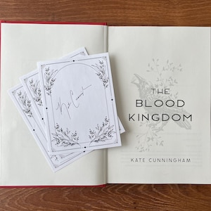 May include: A closed book with a red cover. The title of the book is "The Blood Kingdom" by Kate Cunningham. The book is signed by the author, "Kate Cunningham".
