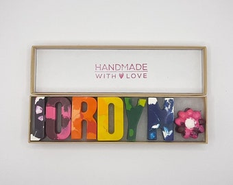 May include: A box of handmade crayons spelling the name "FORDYN" in various colours. The crayons are in the shape of letters and a flower. The box has the text "HANDMADE WITH LOVE".