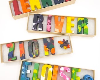 May include: Four boxes of crayons spelling out names: "LENNOX", "RIVER", "ZION", and "ELOISE". The crayons are in a range of colours, including red, orange, yellow, green, blue, and purple. Each name is presented in a rectangular box with a white interior.