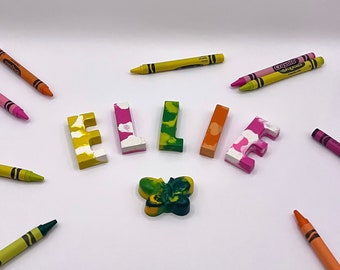 May include: A selection of crayons and wax art spelling "ELLIE". The letters are multi-coloured. A butterfly-shaped crayon is also present, along with individual crayons in shades of orange, yellow, pink, and green, scattered around the letters.