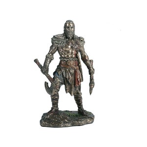 May include: A bronze statue of a warrior holding a double-headed axe and a spear. The warrior is wearing a horned helmet, a fur cloak, and a leather belt with a metal buckle. The statue is standing on a rocky base.
