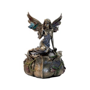 May include: A bronze fairy figurine with iridescent wings sits on a flower-shaped base. The fairy is holding a blue butterfly in her hand. The base is decorated with green leaves and pink flowers.