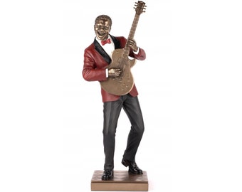 May include: A bronze sculpture of a man playing a guitar. He is wearing a red jacket, black pants, and black shoes. He is smiling and holding the guitar in his left hand. The guitar is gold and has a black pickguard.
