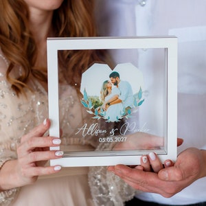 May include: A white frame with a clear glass front holds a photo of a couple. The photo is in the shape of a heart and has a blue and green floral design around it. The text "Allison & Parker 05.05.2027" is printed below the photo.