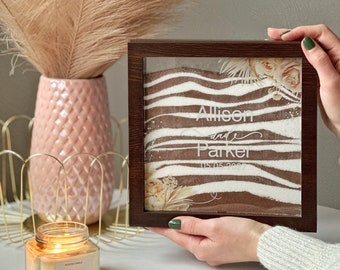 May include: A brown wooden frame with a glass front containing white sand with a white and brown zebra print design. The sand is layered to create a wave effect. The names "Allison and Parker" and the date "05.05.2023" are written in white on the sand. The frame is decorated with dried flowers and leaves.