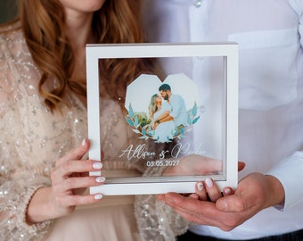 May include: A white frame with a clear glass front holds a photo of a couple. The photo is in the shape of a heart and has a blue and green floral design around it. The text "Allison & Parker 05.05.2027" is printed below the photo.