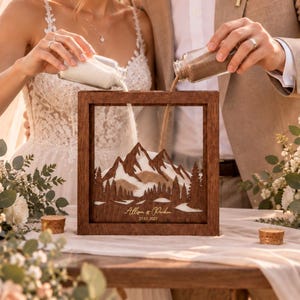 May include: A square wooden sand ceremony frame featuring a laser-cut mountain and forest landscape inside. The frame is engraved with the text Allison & Parker and the date 27.07.2027. Two glass bottles are used to pour white and tan sand into the frame.