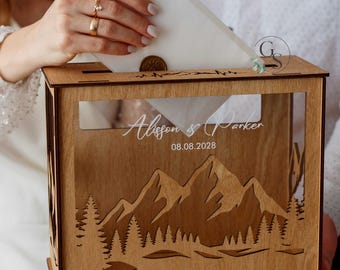 May include: Wooden wedding card box with a mountain and tree design. The box has a clear front panel and the names "Alison & Parker" and the date "08.08.2028" are etched on the front. A person is placing a card into the box.