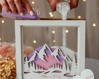 May include: A white framed sand ceremony shadow box with a mountain and tree design. Purple and white sand is being poured into the box. The names "Allison & Parker" and the date "27.07.2027" are printed on the box.