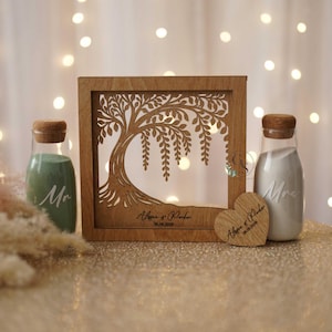 Peut inclure: Un ensemble de cérémonie de mariage en bois avec sable. L'ensemble comprend un cadre en bois avec un motif d'arbre, deux bouteilles en verre avec des bouchons en liège et un cœur en bois. Le cadre est gravé de noms et d'une date. Un flacon contient du sable vert, l'autre blanc.