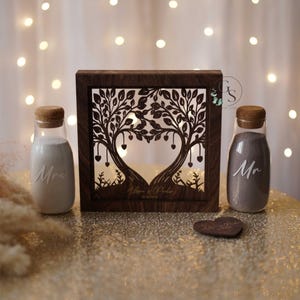 May include: A wooden wedding unity sand ceremony set. The set includes a dark brown wooden frame with a tree design, two glass bottles with cork stoppers, and a heart-shaped wooden piece. The bottles are filled with sand and labeled "Mrs" and "Mr".