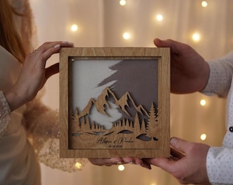 May include: A wooden framed art piece with a mountain and forest design. The artwork features a layered effect with a light and dark color scheme. The frame has the names "Alison & Parker" and the date "08.06.2020" engraved on it.