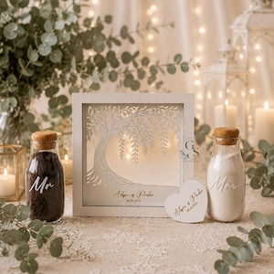 May include: A white square shadow box frame featuring a laser-cut willow tree design, with the text Alison & Parker 08.08.2028. Two glass bottles with cork stoppers, one filled with black sand labeled Mr and one with white sand labeled Mrs.