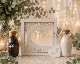 May include: A white square shadow box frame featuring a laser-cut willow tree design, with the text Alison & Parker 08.08.2028. Two glass bottles with cork stoppers, one filled with black sand labeled Mr and one with white sand labeled Mrs.