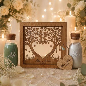 May include: A square wooden frame featuring a laser-cut tree design forming a heart shape in the center, with the text "Alyson & Parker 08.08.2028" engraved at the base. Two glass bottles with cork stoppers contain green and white sand, labeled "Mr" and "Mrs".