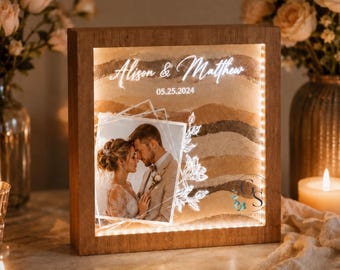 May include: A square wooden light box featuring a sand art display with layered brown tones. The front panel displays a photograph of a couple, the names Alison & Matthew, the date 05.25.2024, and a white floral line drawing. The frame is illuminated.