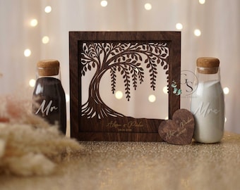 May include: A dark brown wooden wedding keepsake with a tree design, personalized with names and a date. Two glass bottles with cork stoppers, labeled "Mr" and "Mrs", flank the frame. A wooden heart with names and date is also present.