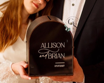 May include: A black wooden wedding card box with a gold arch design. The box has the names "Allison and Brian" and the date "07.07.2027" written on it.
