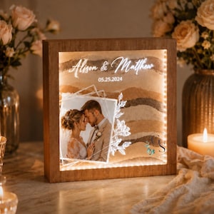 May include: A square wooden light box featuring a sand art display with layered brown tones. The front panel displays a photograph of a couple, the names Alison & Matthew, the date 05.25.2024, and a white floral line drawing. The frame is illuminated.