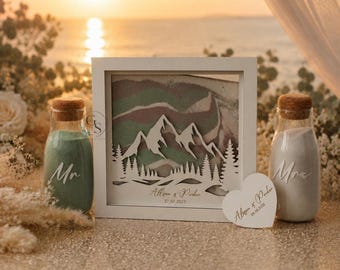 May include: A square white frame containing a layered mountain and forest cutout with colored sand, flanked by two glass bottles with cork stoppers filled with green and white sand, labeled Mr and Mrs, and a heart-shaped tag with names and a date.