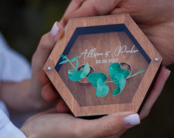 May include: A hexagonal wooden ring box with a clear lid. The box is decorated with a green leafy design and the text "Allison & Parker 05.05.2027".