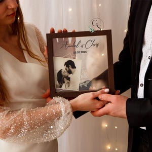 May include: A couple holds a brown wooden frame with a clear glass front. The frame contains a black and white photo of a couple kissing. The frame has the text "Annabel & Chris 05.05.2023" printed on it.