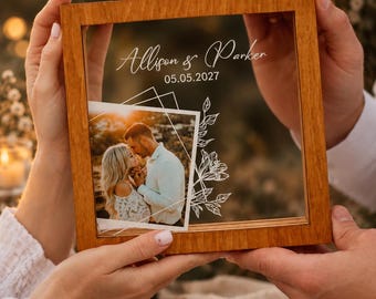 May include: A square wooden frame containing a clear pane with the white printed text Allison & Parker and the date 05.05.2027. A photograph of a couple is attached to the glass, accented by a white line drawing of geometric shapes and flowers.