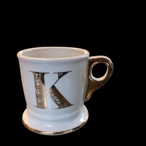 May include: White ceramic mug with a gold letter K on the front. The mug has a gold rim and a gold handle.