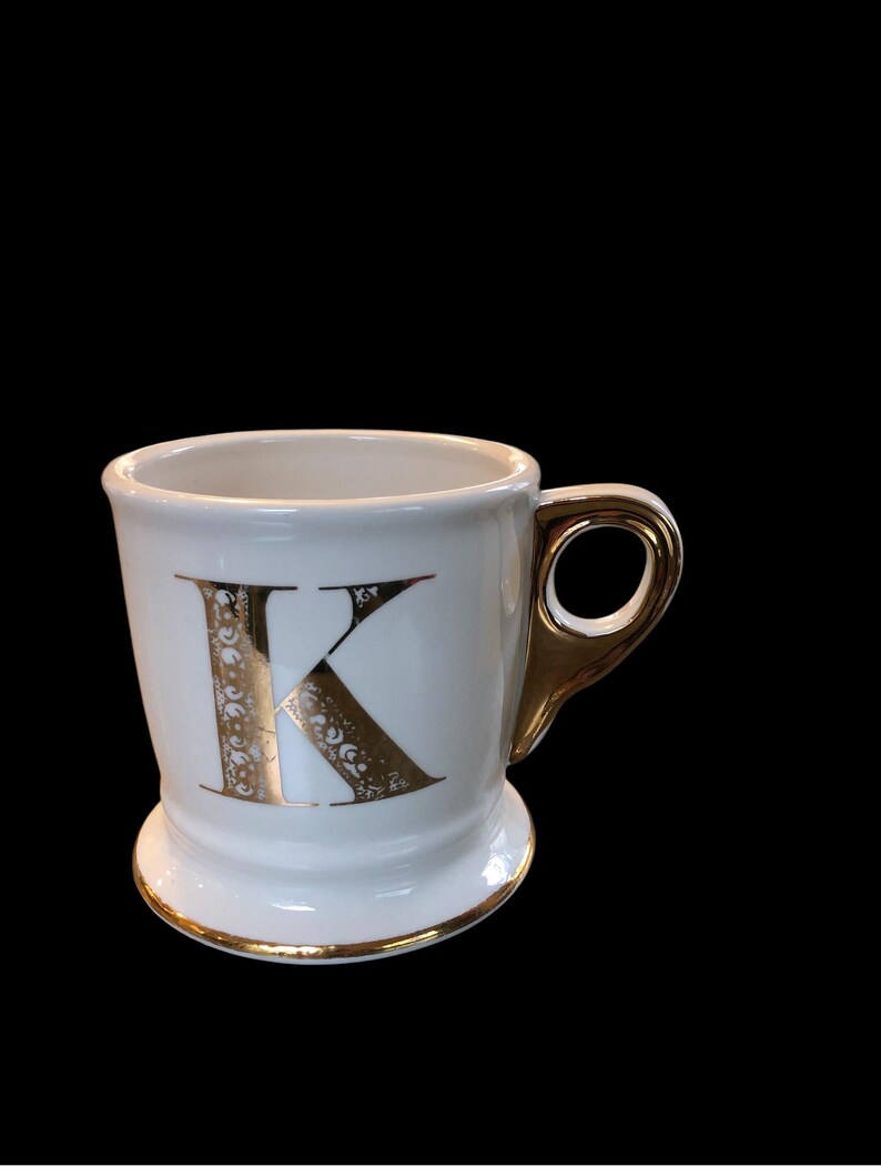 May include: White ceramic mug with a gold letter K on the front. The mug has a gold rim and a gold handle.