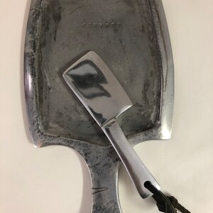 May include: A silver cheese board with a small knife attached. The board has a rounded rectangular shape with a handle on the bottom. The board is marked with the word "Gourmet".
