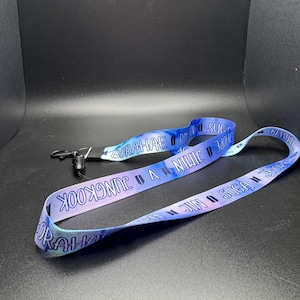 May include: A blue and purple lanyard with the words "JUNGKOOK", "SUGA", "JIN", "JIMIN", "V", "HOPE", and "BORAHAE" printed on it. The lanyard has a black clip and a tie-dye pattern.
