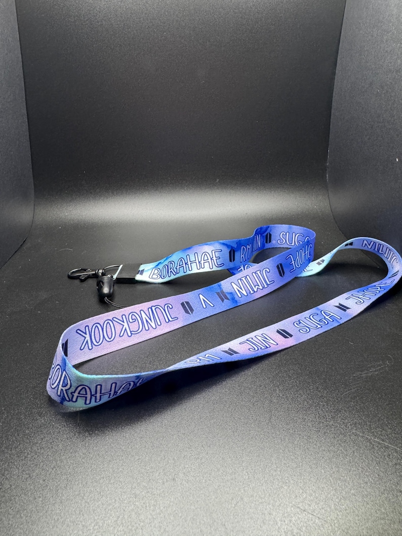 May include: A blue and purple lanyard with the words "JUNGKOOK", "SUGA", "JIN", "JIMIN", "V", "HOPE", and "BORAHAE" printed on it. The lanyard has a black clip and a tie-dye pattern.