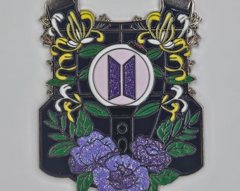May include: A black enamel pin with a purple and white stylized logo, surrounded by yellow and white flowers and purple flowers with glitter accents.
