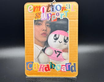 May include: A small wooden easel displaying a cardboard picture frame with a colorful "Emotional Support Cardboard" title. The frame contains a photo of a person holding a pink and white plush toy.