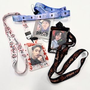 May include: A collection of lanyards and ID holders featuring images of K-pop stars. The lanyards are in various colors, including white, blue, and black, with printed text and graphics. The ID holders display photos and the word "BIASED".