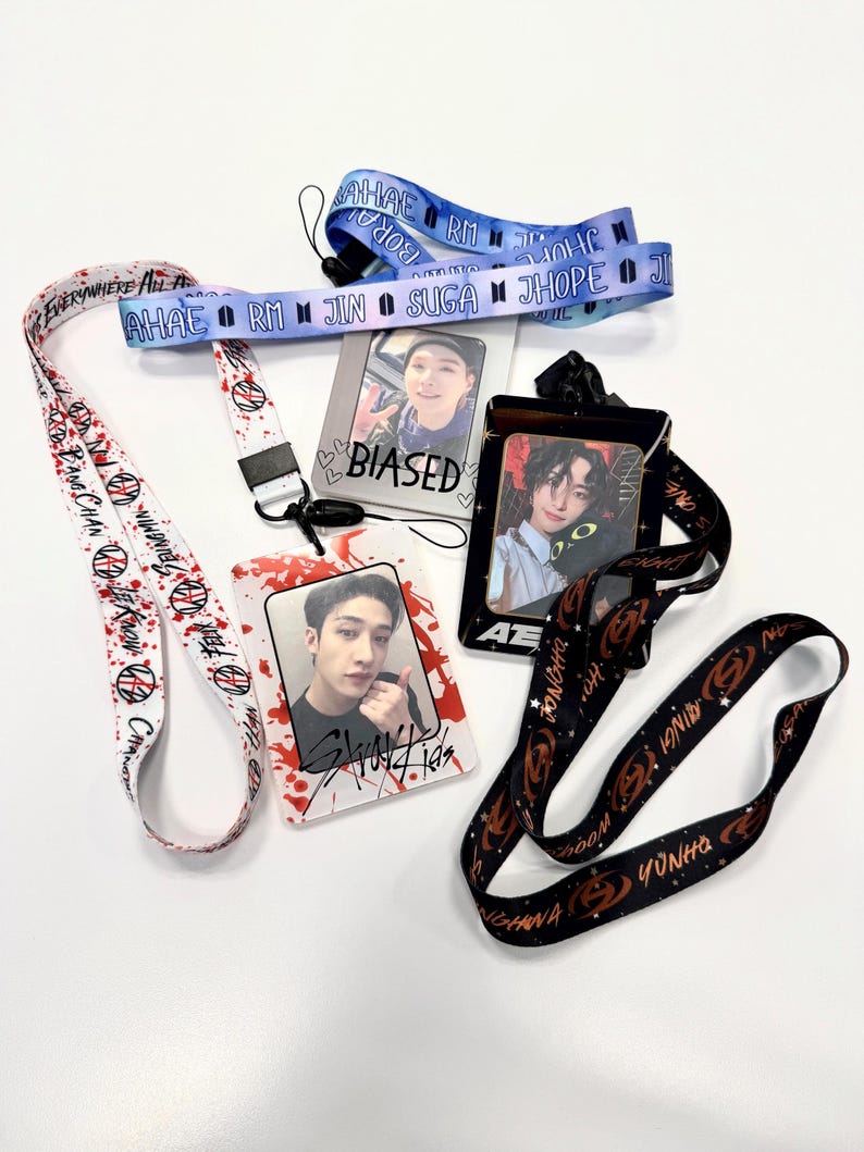 May include: A collection of lanyards and ID holders featuring images of K-pop stars. The lanyards are in various colors, including white, blue, and black, with printed text and graphics. The ID holders display photos and the word "BIASED".